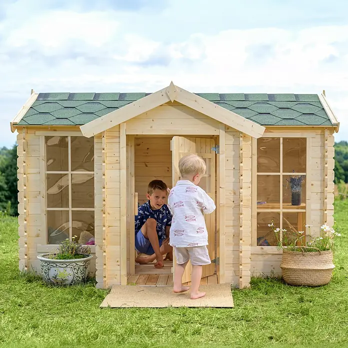 Timbela Spielhaus - L x B x H: 187 x 241 x 151 cm, Holz, Natur - Zwei Kinder spielen in einem hellen Holzspielhaus mit grünem Dach, das mit Blumenkästen verziert ist. Das Spielhaus steht auf einer grünen Wiese.