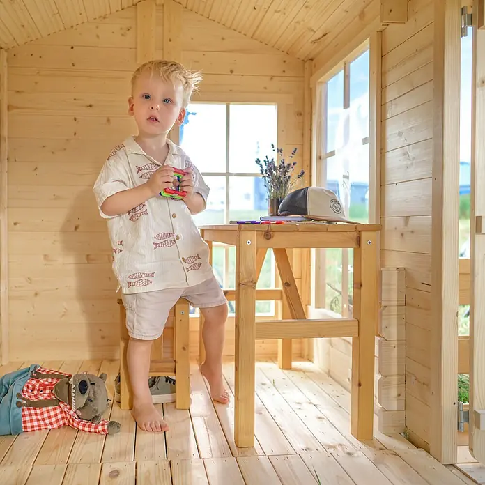 Timbela Spielhaus - L x B x H: 187 x 241 x 151 cm, Holz, Natur - Ein niedlicher Junge spielt in einem Holzhaus mit einem Spielzeug in der Hand, während er an einem Holztisch steht.