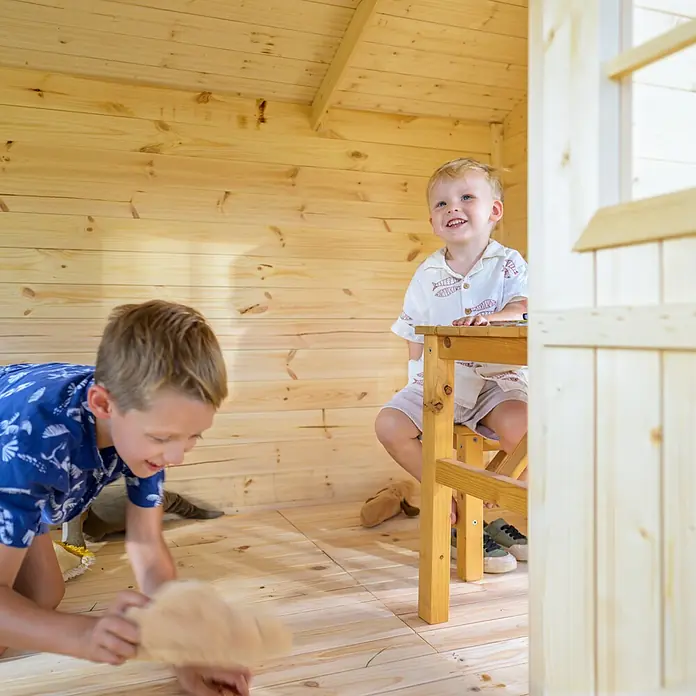 Timbela Spielhaus - L x B x H: 187 x 241 x 151 cm, Holz, Natur - Ein helles, hölzernes Kinderspielhaus mit zwei spielenden Jungen, einer sitzt am Tisch, der andere spielt auf dem Boden.