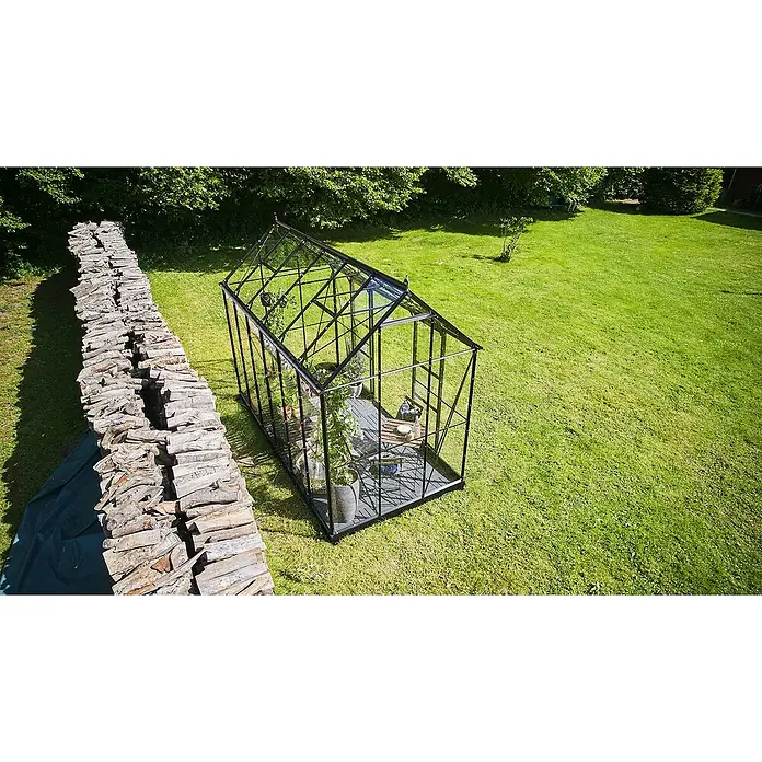 Halls Gewächshaus Icon Planthouse - Außenmaß inkl. Dachüberstand (B x T): 379,1 x 192,5 cm, Material Seitenwand: Glas, 3 mm, Schwarz - Ein Gewächshaus mit Pflanzen steht auf einer Rasenfläche neben einem Stapel Holz. Es bietet Schutz und verlängert die Gartensaison.