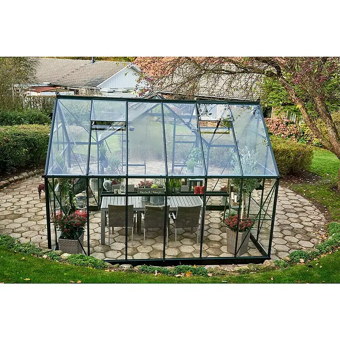 Halls Gewächshaus Icon Gardenhouse - Außenmaß inkl. Dachüberstand (B x T): 383,9 x 381,5 cm, Material Seitenwand: Glas, 3 mm, Grün - Das Bild zeigt ein Gewächshaus mit einem Tisch und Stühlen darin, umgeben von einem Garten.