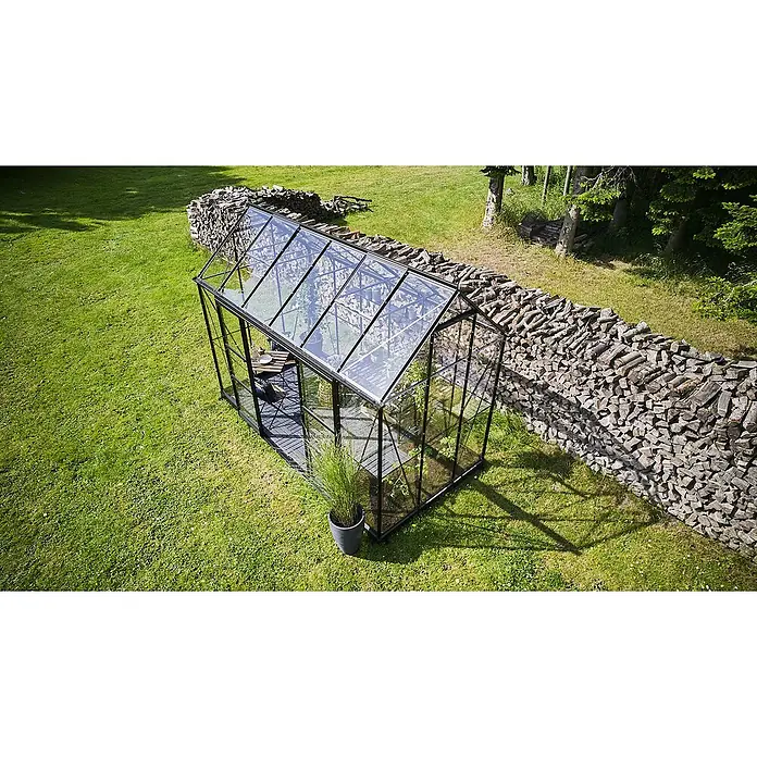 Halls Gewächshaus Icon Planthouse - Außenmaß inkl. Dachüberstand (B x T): 379,1 x 192,5 cm, Material Seitenwand: Glas, 3 mm, Schwarz - Ein modernes Gewächshaus mit Glaswänden und schwarzem Rahmen steht auf einer grünen Wiese neben einem Stapel Brennholz.