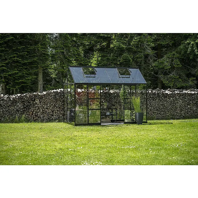 Halls Gewächshaus Icon Planthouse - Außenmaß inkl. Dachüberstand (B x T): 379,1 x 192,5 cm, Material Seitenwand: Glas, 3 mm, Schwarz - Das Gewächshaus mit schwarzem Rahmen steht auf einer grünen Wiese vor einer Holzstapelwand und bietet Platz für Pflanzen und Gemüse.