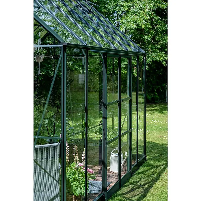 Halls Gewächshaus Icon Planthouse - Außenmaß inkl. Dachüberstand (B x T): 379,1 x 254,7 cm, Material Seitenwand: Glas, 3 mm, Grün - Nahaufnahme eines Gewächshauses mit Pflanzen, Werkzeugen und einem Stuhl, das eine ruhige Gartenszene zeigt.