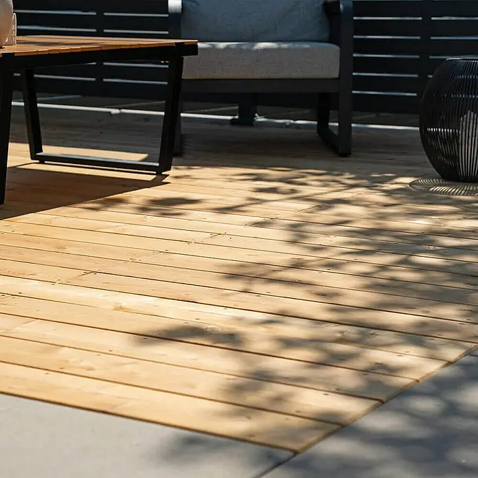Home Deluxe Terrassenfliese ARIS Lärche 40 m² - Braun, Matt - Nahaufnahme eines Holzdecks mit einer dunklen Tischgarnitur und einem Sessel mit grauen Kissen im Hintergrund.