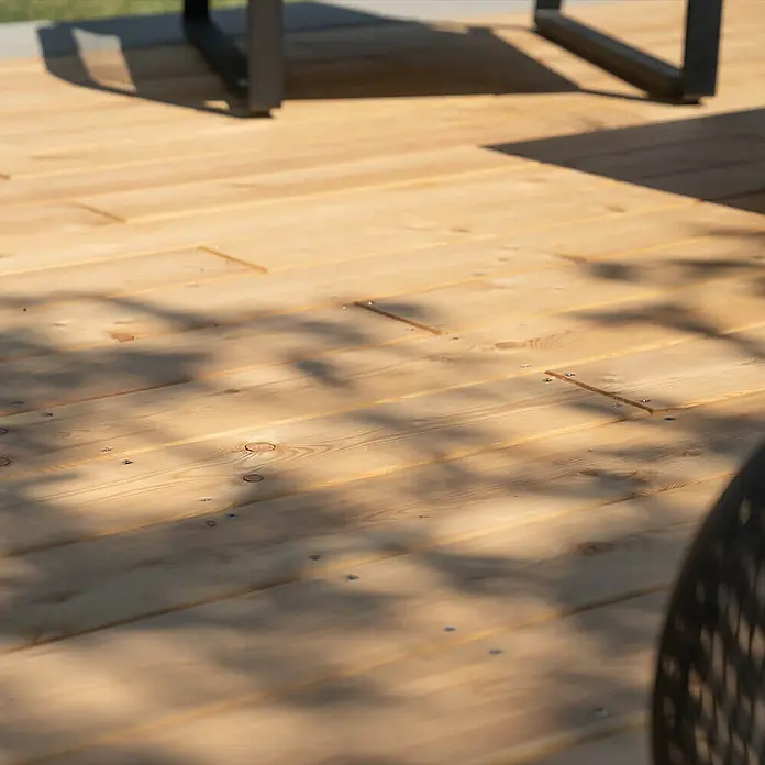 Home Deluxe Terrassenfliese ARIS Lärche 50 m² - Braun, Matt - Nahaufnahme einer hellen Holzterrasse mit Tischbeinen und Schatten. Das natürliche Holz und die subtilen Schatten sorgen für eine warme Atmosphäre.