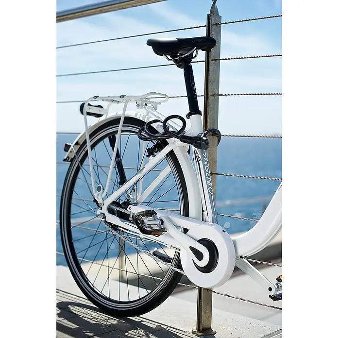 Abus Spiralkabelschloss Booster 6512K - Länge: 180 cm - Weißes Argemo-Fahrrad, angeschlossen an ein Geländer mit Blick aufs Meer. Das Rad hat einen Gepäckträger, Kettenschutz und ein Schloss.