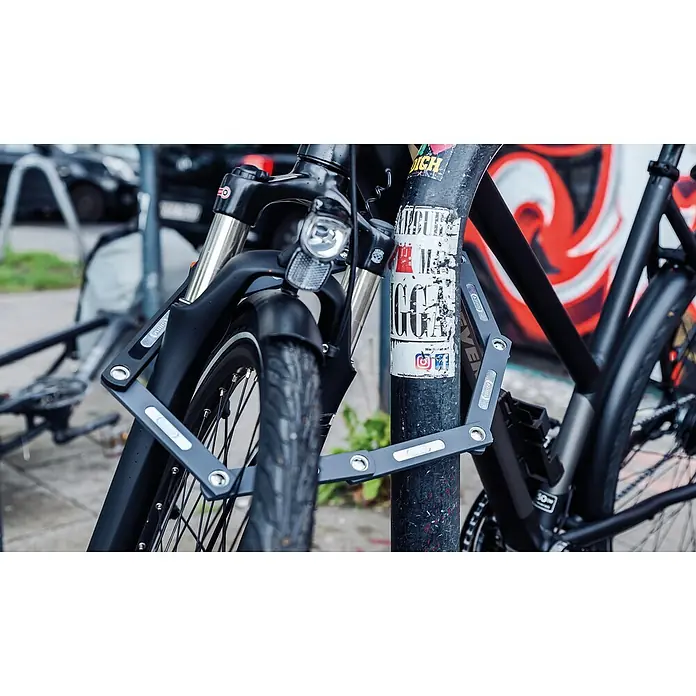 Abus Faltschloss 5700K - Länge: 100 cm - Das ABUS Faltschloss sichert ein Fahrrad an einem Pfosten, wobei die robuste Konstruktion und die praktische Faltbarkeit des Schlosses hervorstechen.