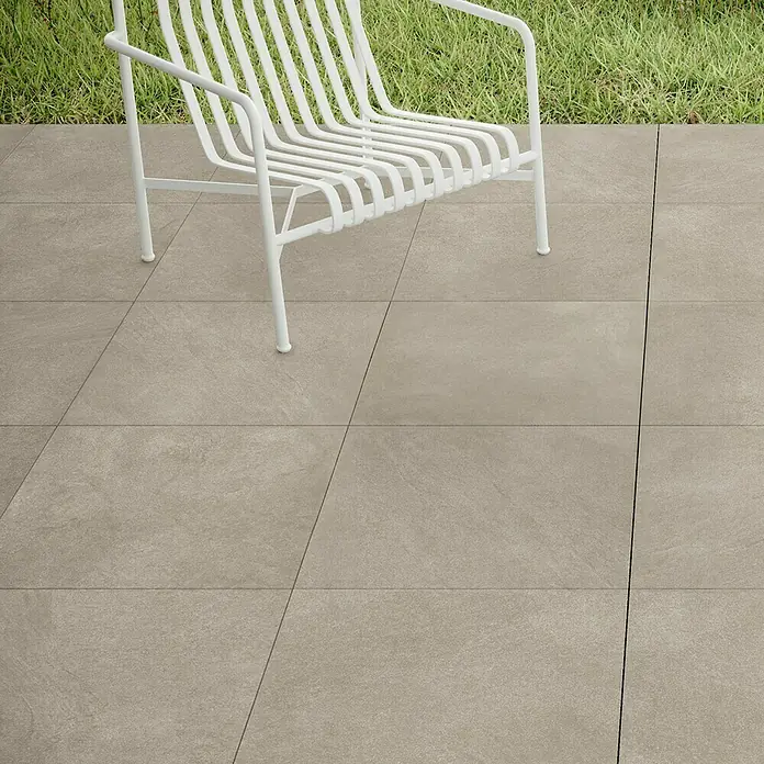 Terrassenfliese Stoneline - 60 x 60 x 2 cm, Beige/Grau, Matt - Nahaufnahme von hellgrauen Steinzeugfliesen auf einer Terrasse mit einem weißen Stuhl. Die Fliesen haben eine matte Oberfläche.