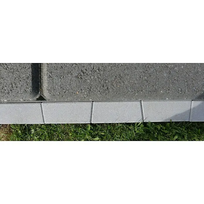 Christoph Mähstein Trapez - 24 x 10 x 4,2 cm, Silbergrau, Beton - Nahaufnahme von grauen Pflastersteinen, die einen Gehwegrand bilden, mit Gras im Vordergrund. Die Steine sind sauber und gut verarbeitet.