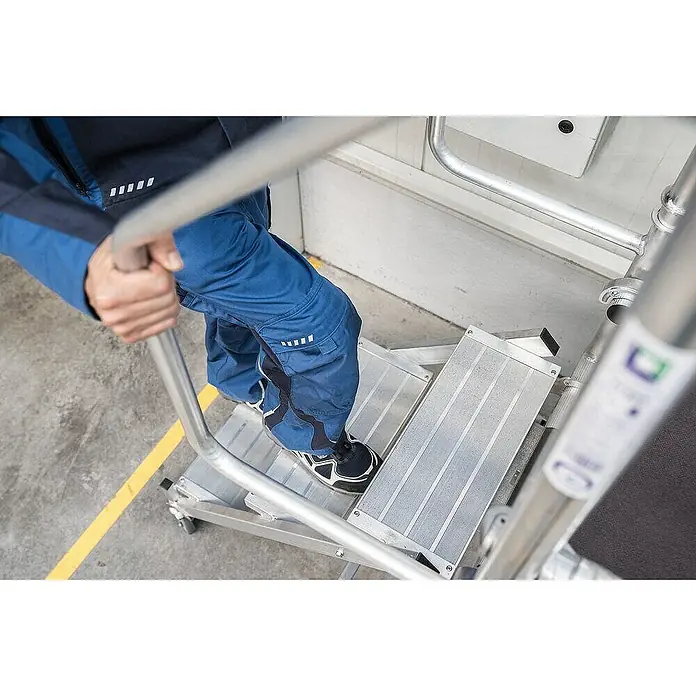 Proregal Montagegerüst mit Treppenzustieg -  - Person besteigt eine silberne, mobile Arbeitsplattform mit rutschfesten Metallstufen.