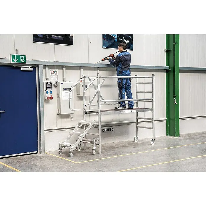 Proregal Montagegerüst mit Treppenzustieg -  - Silbernes, mobiles Aluminium-Rollgerüst mit Treppe und Rädern, von einem Mann bei Arbeiten an einer Wand genutzt.