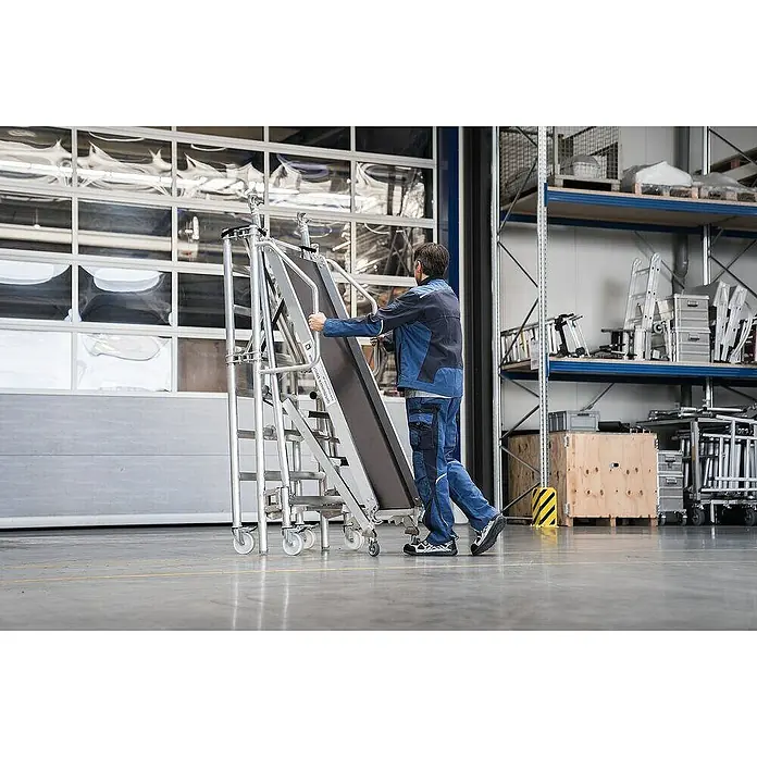 Proregal Montagegerüst mit Treppenzustieg -  - Mann bewegt eine silberne, mobile Arbeitsbühne aus Aluminium mit schwarzen Stufen und Rollen in einer Halle.