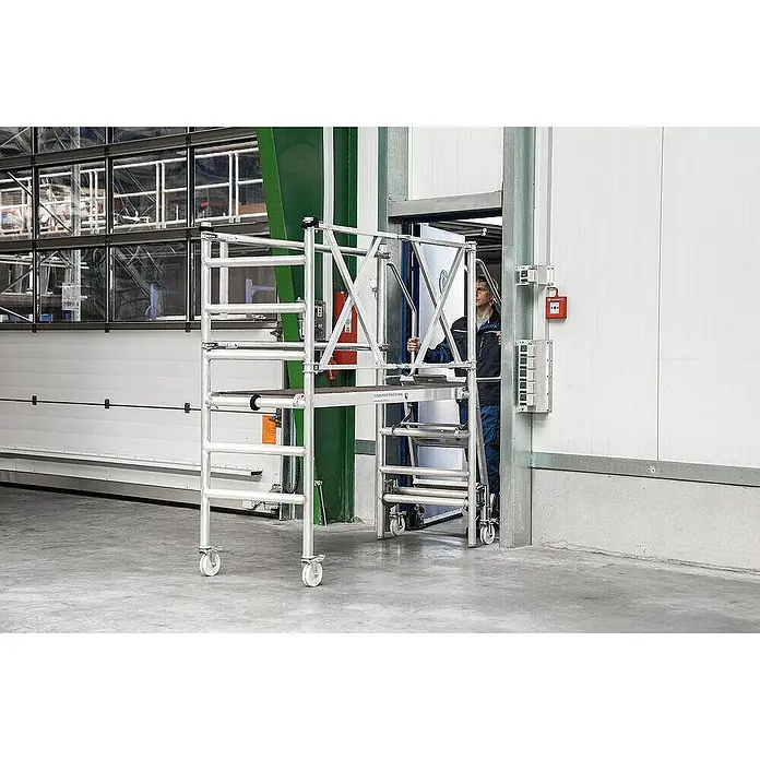 Proregal Montagegerüst mit Treppenzustieg -  - Mobiles Aluminiumgerüst mit Arbeiter auf der Plattform, der es durch einen Türrahmen in einem Industriegebäude bewegt.