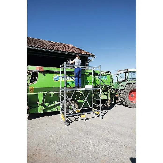 Proregal Klappgerüst mit Ausbaueinheit -  - Mobiles, silberfarbenes Aluminium-Rollgerüst mit Rädern, von einer Person bei Arbeiten in der Höhe an landwirtschaftlichen Maschinen genutzt.