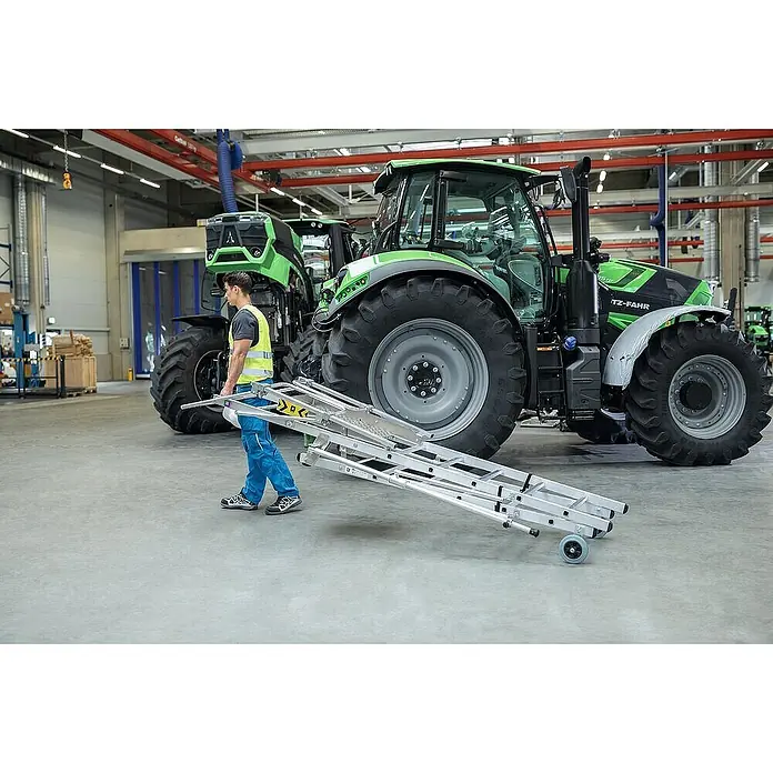 Proregal Stehleiter Plattformleiter Flexstep - Anzahl Tritte: 6 Stk., Belastbarkeit: 150 kg, Aluminium - Mann trägt eine silberne Mehrzweckleiter mit Rollen, was ihre Portabilität in einer Industrieumgebung hervorhebt.