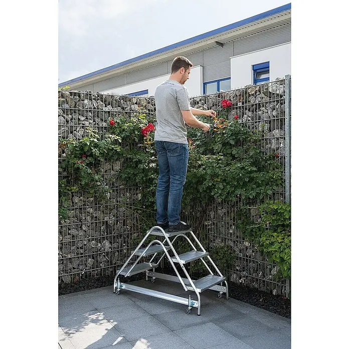 Proregal Treppenpodest Arbeitspodest beidseitig begehbar - Anzahl Tritte: 8 Stk., Arbeitshöhe: 3.000 mm, Belastbarkeit: 150 kg, Aluminium - Mann auf einer silbernen Aluminium-Plattformleiter mit mehreren Stufen, der Rosen im Garten schneidet.