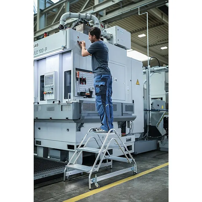Proregal Treppenpodest Arbeitspodest beidseitig begehbar - Anzahl Tritte: 8 Stk., Arbeitshöhe: 3.000 mm, Belastbarkeit: 150 kg, Aluminium - Silberne, fahrbare Arbeitsplattformleiter aus Aluminium, von einem Arbeiter an Industriemaschinen genutzt.