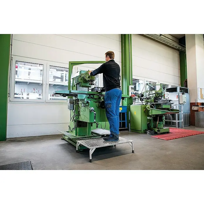 Proregal Treppenpodest Arbeitspodest mit geräumiger Plattform - Arbeitshöhe: 2.250 mm, Belastbarkeit: 150 kg, Aluminium - Trittschemel aus silbernem Riffelblech, von einem Arbeiter zum Erreichen von Maschinen genutzt.