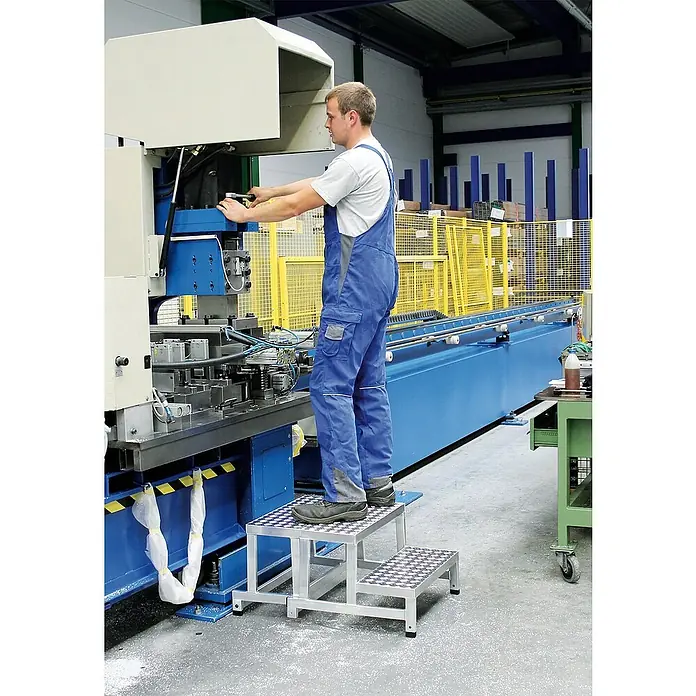 Proregal Treppenpodest Arbeitspodest modular - Anzahl Tritte: 3 Stk., Arbeitshöhe: 2.600 mm, Belastbarkeit: 150 kg, Aluminium - Zweistufige silberne Aluminium-Arbeitsplattform mit rutschfesten Trittflächen, von einem Arbeiter an Maschinen verwendet.