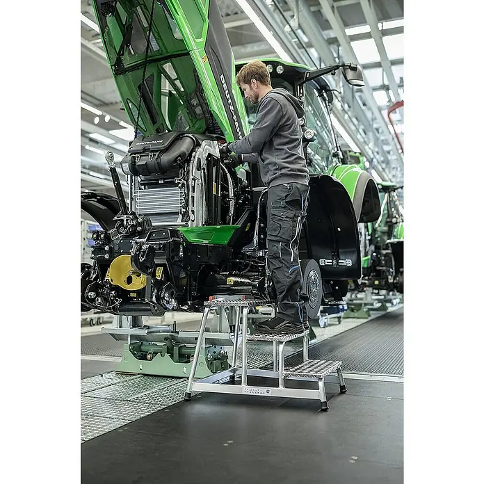 Proregal Treppenpodest Arbeitspodest starr mit 1 Stufe - Anzahl Tritte: 1 Stk., Arbeitshöhe: 2.200 mm, Belastbarkeit: 150 kg, Aluminium - Ein Arbeiter steht auf einem silbernen Aluminium-Trittschemel, um Bauteile an einem großen grünen Traktor zu erreichen.