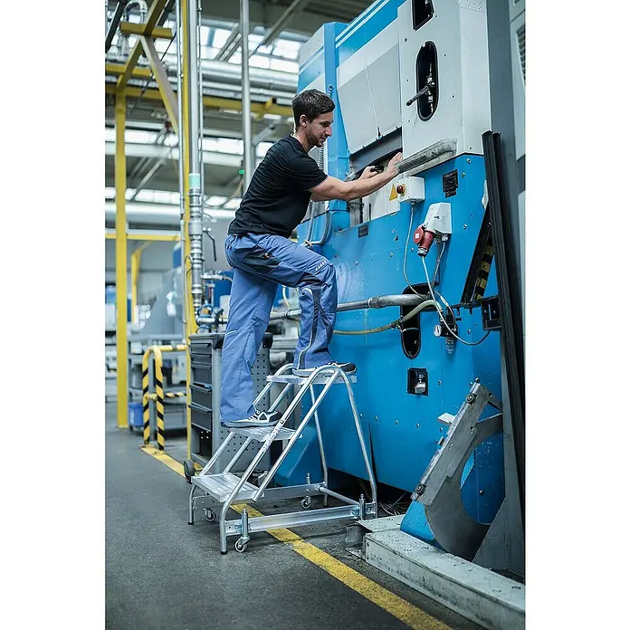 Proregal Treppenpodest Arbeitspodest einseitig begehbar - Anzahl Tritte: 4 Stk., Arbeitshöhe: 3.000 mm, Belastbarkeit: 150 kg, Aluminium - Silberne Aluminium-mobile Plattformtreppe, von einem Mann zum Zugang zu einer Maschine in einer Industrieumgebung genutzt.