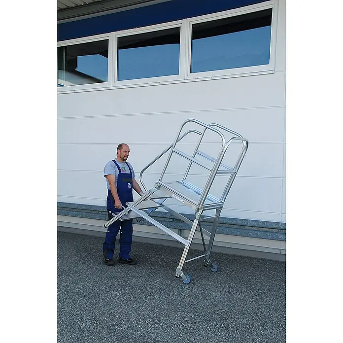 Proregal Treppenpodest einseitig begehbar - Anzahl Tritte: 4 Stk., Arbeitshöhe: 3.000 mm, Belastbarkeit: 150 kg, Aluminium - Mann bewegt eine silberne mobile Arbeitsplattform aus Aluminium mit Handläufen und Rädern.