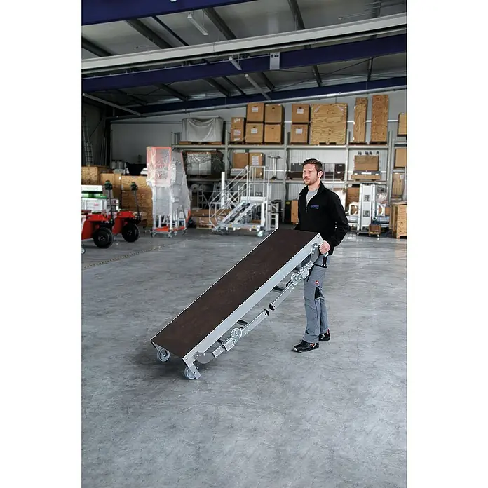 Proregal Treppenpodest Maschinenpodest höhenverstellbar - Arbeitshöhe: 2.750 mm, Belastbarkeit: 300 kg, Aluminium - Mann schiebt mobiles Förderband mit silbernem Gestell und dunklem Gurt in einer Lagerhalle.