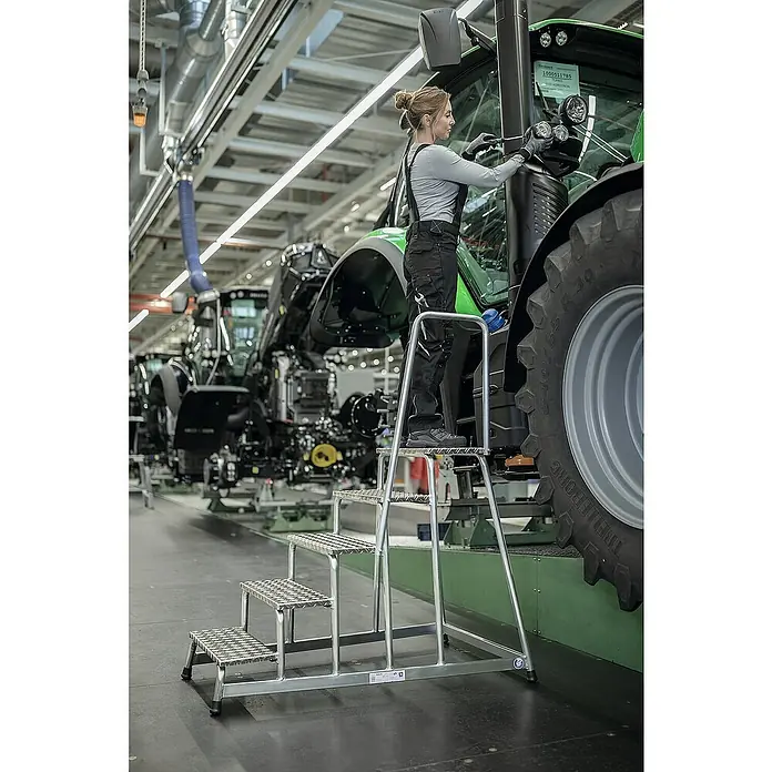 Proregal Treppenpodest Arbeitspodest starr mit 5 Stufen - Anzahl Tritte: 5 Stk., Arbeitshöhe: 3.000 mm, Belastbarkeit: 150 kg, Aluminium - Silberne Aluminium-Industriestufen, die von einem Arbeiter benutzt werden, um einen großen Traktor in einer Fabrik zu erreichen. Bietet sicheren Zugang.