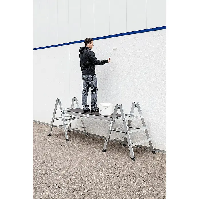 Proregal Doppel-Klapptritt beidseitig begehbar - Anzahl Tritte: 4 Stk., Arbeitshöhe: 2.990 mm, Belastbarkeit: 150 kg, Aluminium - Aluminium-Arbeitsplattform mit dunkler Oberfläche, genutzt von einem Mann, der eine weiße Wand streicht. Ein Eimer steht ebenfalls auf der Plattform.