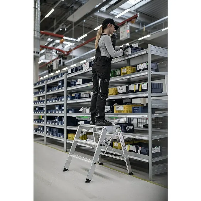Proregal Klapptritt einseitig begehbar - Anzahl Tritte: 2 Stk., Arbeitshöhe: 2.700 mm, Belastbarkeit: 150 kg, Aluminium - Silberne Aluminium-Klappleiter, die von einer Person zum Erreichen von Gegenständen in hohen Lagerregalen verwendet wird.
