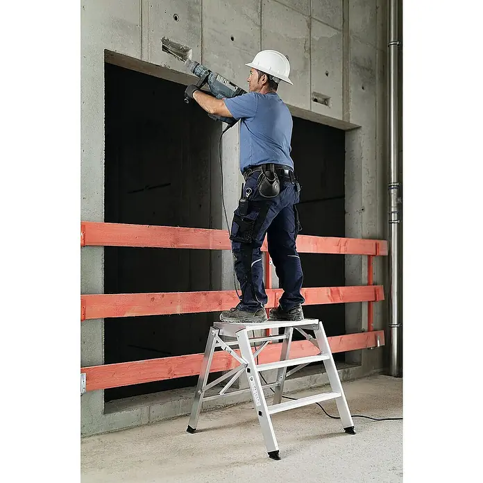 Proregal Klapptritt einseitig begehbar - Anzahl Tritte: 3 Stk., Arbeitshöhe: 3.000 mm, Belastbarkeit: 150 kg, Aluminium - Silberner Klapptritt, von einem Bauarbeiter zum Bohren an einer Wand genutzt.