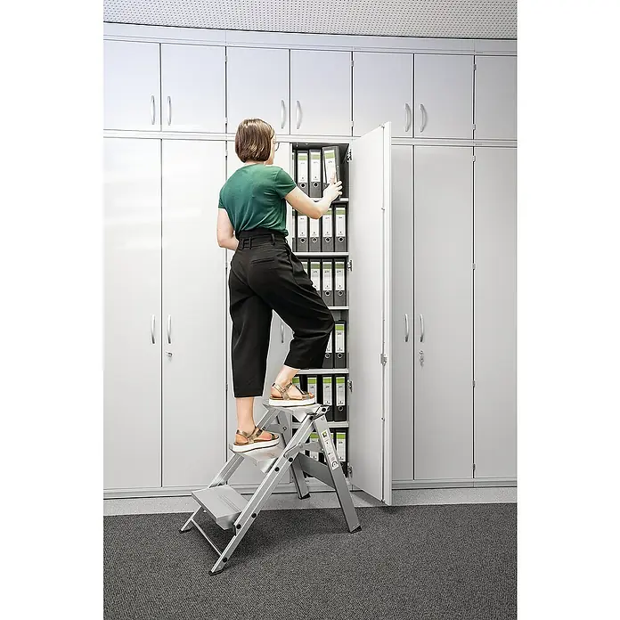 Proregal Klapptritt mit 3 Stufen - Anzahl Tritte: 3 Stk., Arbeitshöhe: 2.700 mm, Belastbarkeit: 150 kg, Aluminium - Alu-Klapptritt, der einer Frau hilft, Akten in einem hohen Schrank zu erreichen.