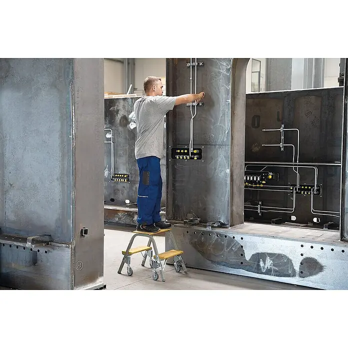 Proregal Doppel-Stehleiter mit Lenkrollen - Anzahl Tritte: 4 Stk., Arbeitshöhe: 2.400 mm, Belastbarkeit: 150 kg, Stahl - Mann auf rollendem Tritthocker mit Metallrahmen und gelben Holzstufen, der Teile an Industriemaschinen montiert.