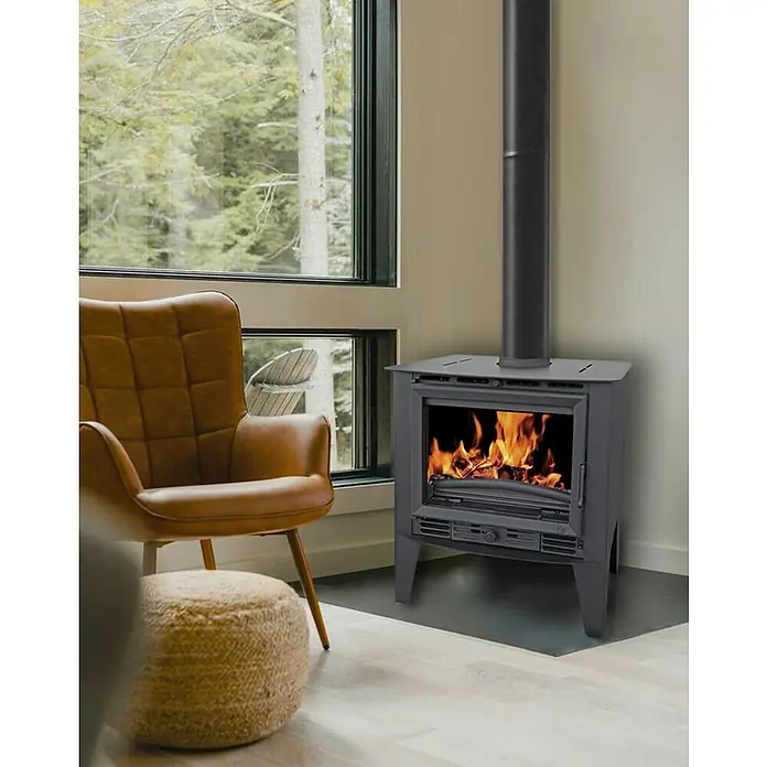 Invicta Estufa de leña Siena - 8 kW, Volumen de calefacción: 240 m³, sin ventilador, Antracita - Acogedora sala de estar con una estufa de leña moderna y un sillón cómodo. La estufa proporciona calor y ambiente.