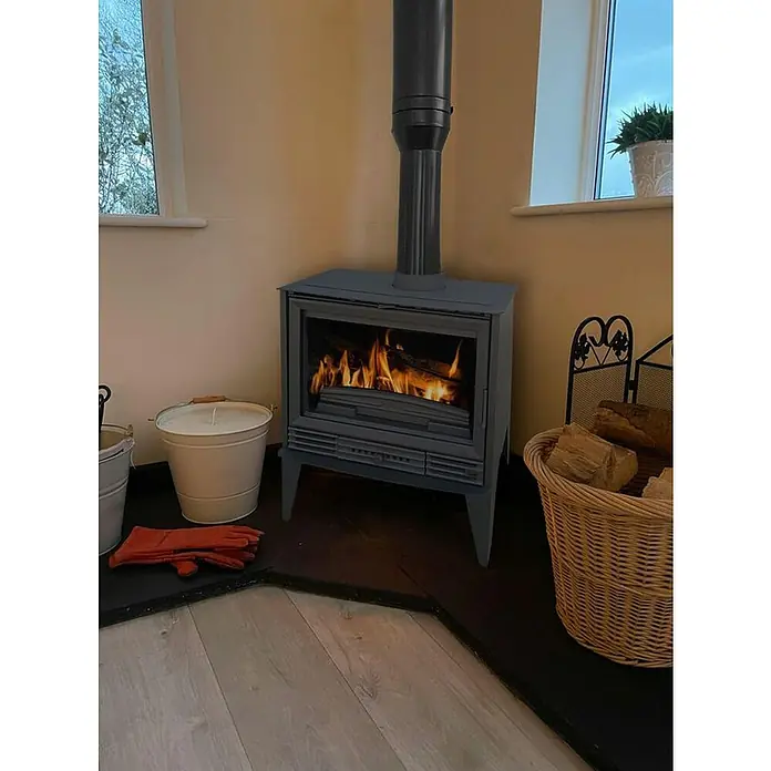 Invicta Estufa de leña Padua sin ventiladores - 10 kW, Volumen de calefacción: 300 m³, sin ventiladores, Negro - Estufa de leña oscura con leña ardiendo, rodeada de accesorios rústicos como un cubo de leña y una cesta.