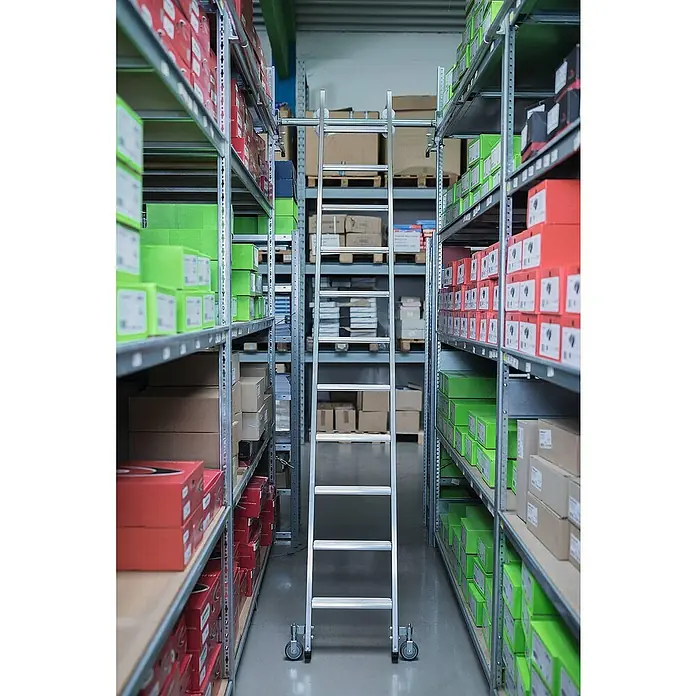 Proregal Anlegeleiter Regalleiter fahrbar - Anzahl Tritte: 10 Stk., Arbeitshöhe: 3.590 mm, Belastbarkeit: 150 kg, Aluminium - Silberne Rollleiter in einem Lagergang zwischen Regalen voller Kartons. Zum Erreichen hoher Regale.
