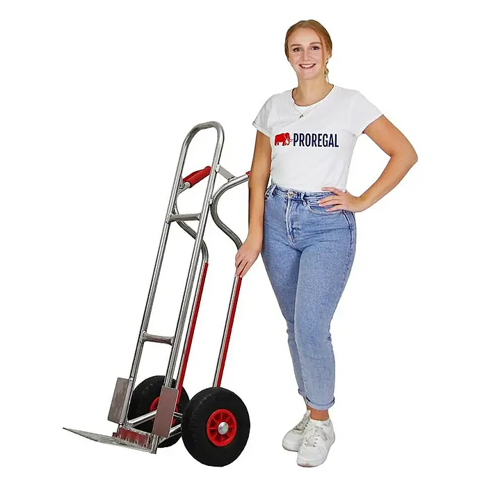 Proregal Sackkarre RAGNAR mit klappbarer Schaufel & PU-Bereifung - Aluminium, Traglast: 250 kg - Junge Frau neben einer silbernen Sackkarre mit roten Griffen und schwarzen Luftreifen. Bereit für schwere Transporte.
