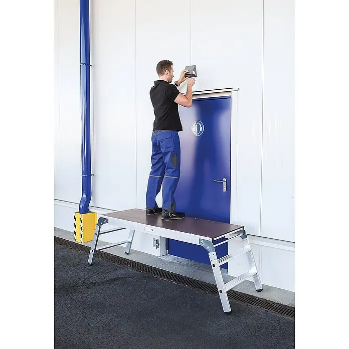 Proregal Treppenpodest Arbeitsdiele klappbar - Arbeitshöhe: 2.700 mm, Belastbarkeit: 300 kg, Aluminium - Mann steht auf einer silbernen Aluminium-Arbeitsplattform mit dunkler Oberseite und montiert einen Gegenstand über einer blauen Tür.