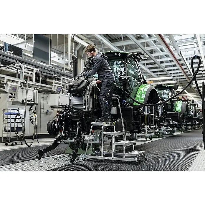 Proregal Treppenpodest Arbeitspodest starr mit 2 Stufen - Anzahl Tritte: 2 Stk., Belastbarkeit: 150 kg, Aluminium - Silberfarbener Alu-Rolltritt mit mehreren Stufen, von einem Arbeiter bei der Traktormontage in einer Fabrik genutzt.