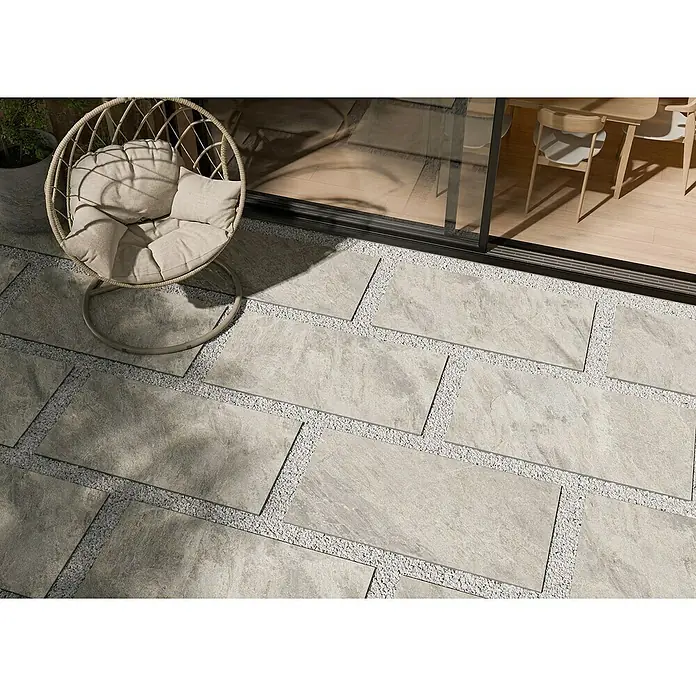Terrassenfliese Homecraft - 59,7 x 119,7 x 2 cm, Grey, Matt - Eine elegante graue Terrassenfliese mit Kiesakzenten und einem modernen Sessel, ideal für stilvolle Außenbereiche.