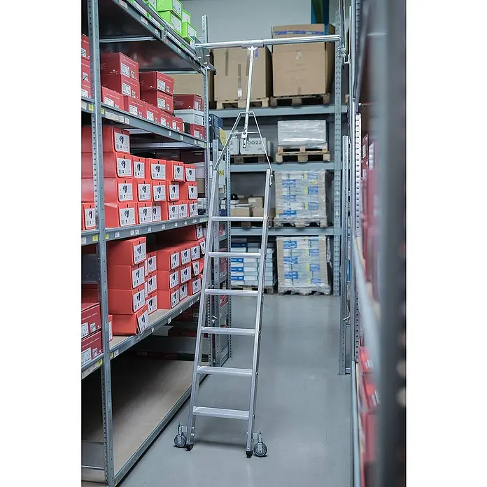 Proregal Anlegeleiter Regalleiter drehabr mit 11 Stufen - Anzahl Tritte: 11 Stk., Arbeitshöhe: 4.610 mm, Belastbarkeit: 150 kg, Aluminium - Silberne Aluminium-Rollleiter mit Rädern, an einer oberen Schiene in einem Lagergang mit Regalen befestigt.