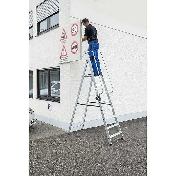 Proregal Stehleiter mit 8 Stufen - Anzahl Tritte: 8 Stk., Belastbarkeit: 150 kg, Aluminium - Mann auf einer Aluminium-Arbeitsplattform montiert ein Schild an einem Gebäude. Die silberne Leiter hat einen Sicherheitsgeländer.