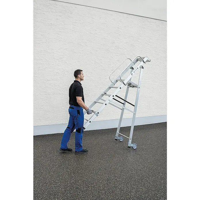Proregal Stehleiter mit Rollen und Griff - Anzahl Tritte: 8 Stk., Belastbarkeit: 150 kg, Aluminium - Mann bewegt eine silberne Aluminium-Arbeitsplattform mit integrierten Stufen und Handläufen, ausgestattet mit Rollen zum Transport.