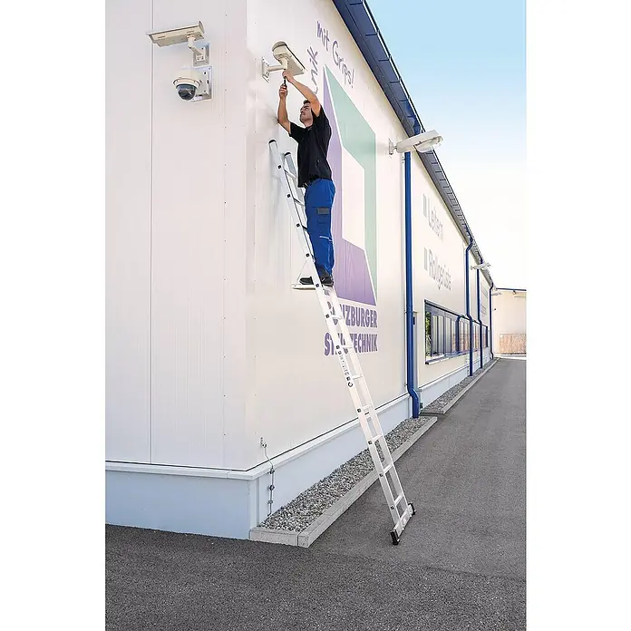 Proregal Anlegeleiter mit Standard-Traverse - Anzahl Tritte: 20 Stk., Arbeitshöhe: 6.900 mm, Belastbarkeit: 150 kg, Aluminium - Mann auf einer silbernen Leiter installiert eine Überwachungskamera an einem Gebäude. Die robuste Leiter bietet sicheren erhöhten Zugang.
