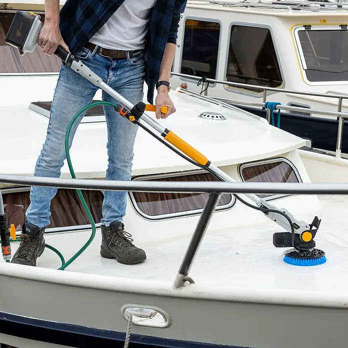 Batavia Maxxpack Akku-Reinigungsbürste Twin Brush V1 - 18 V, 1 Akku, 4 Ah - Eine Person reinigt ein Bootsdeck mit einer rotierenden Bürste an einem Teleskopstiel. Die Bürste hat blaue Borsten und einen gelben Kopf.