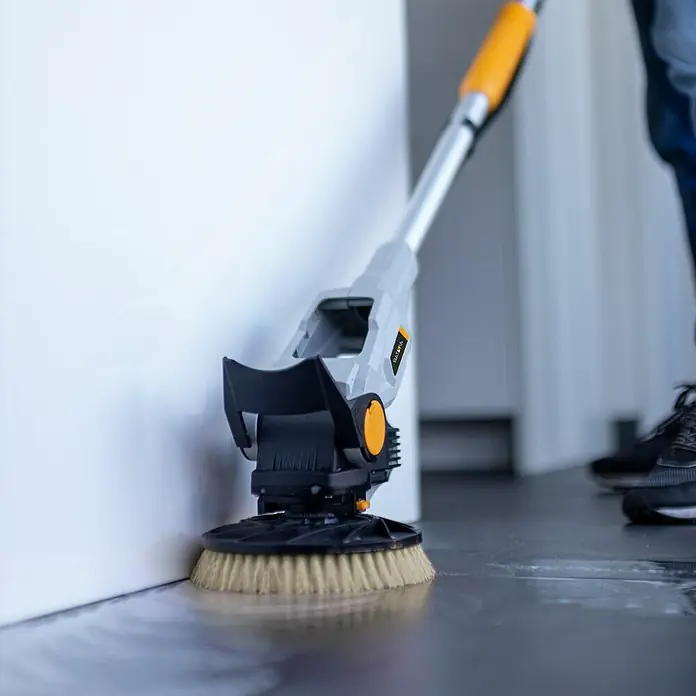Batavia Maxxpack Reinigungsbürste - Material Borsten: Nylon, Passend für: Batavia Akku-Reinigungsbürste Twin Brush - Ein Akku-Bodenwischer mit Bürstenkopf wird an einer Wand verwendet, um den Boden zu reinigen. Das Gerät hat einen gelben Griff und einen grauen Korpus.