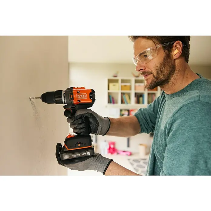 Black+Decker 18V PowerConnect Taladro atornillador percutor de batería BLD683XN-XJ - 18 V, Sin batería, 50 Nm, Número máx. de impactos: 28.800 /min - Un hombre taladra una pared con un taladro inalámbrico Black+Decker de 18 V, con gafas de seguridad y guantes.