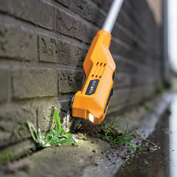 Batavia Fixxpack Unkrautbürste Fixxbrush - 12 V, Li-Ionen, Ohne Akku - Nahaufnahme eines orangefarbenen Batavia-Unkrautentferners, der an einer Ziegelmauer Unkraut entfernt und Licht auf die Arbeit wirft.