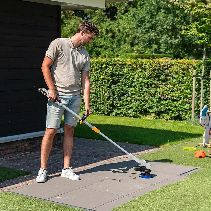 Batavia Maxxpack Akku-Reinigungsbürste Twin Brush Compact - 18 V, Ohne Akku - Mann benutzt eine Fugenkratzbürste, um Unkraut von einer Terrasse zu entfernen. Das Gerät hat eine lange Stange für eine bequeme Nutzung.